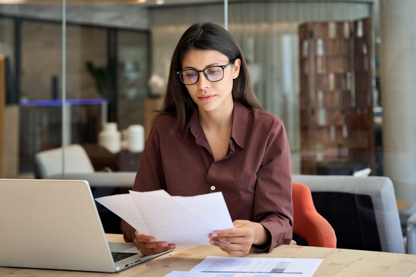 A woman sits at a table with her laptop while reading a report from paper she is holding in her hands